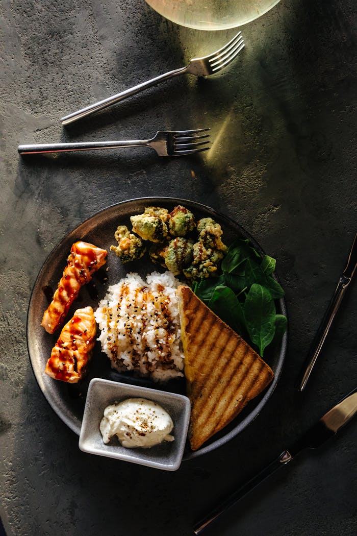 A gourmet dish featuring grilled salmon, rice, greens, and toast on a dark table setting.